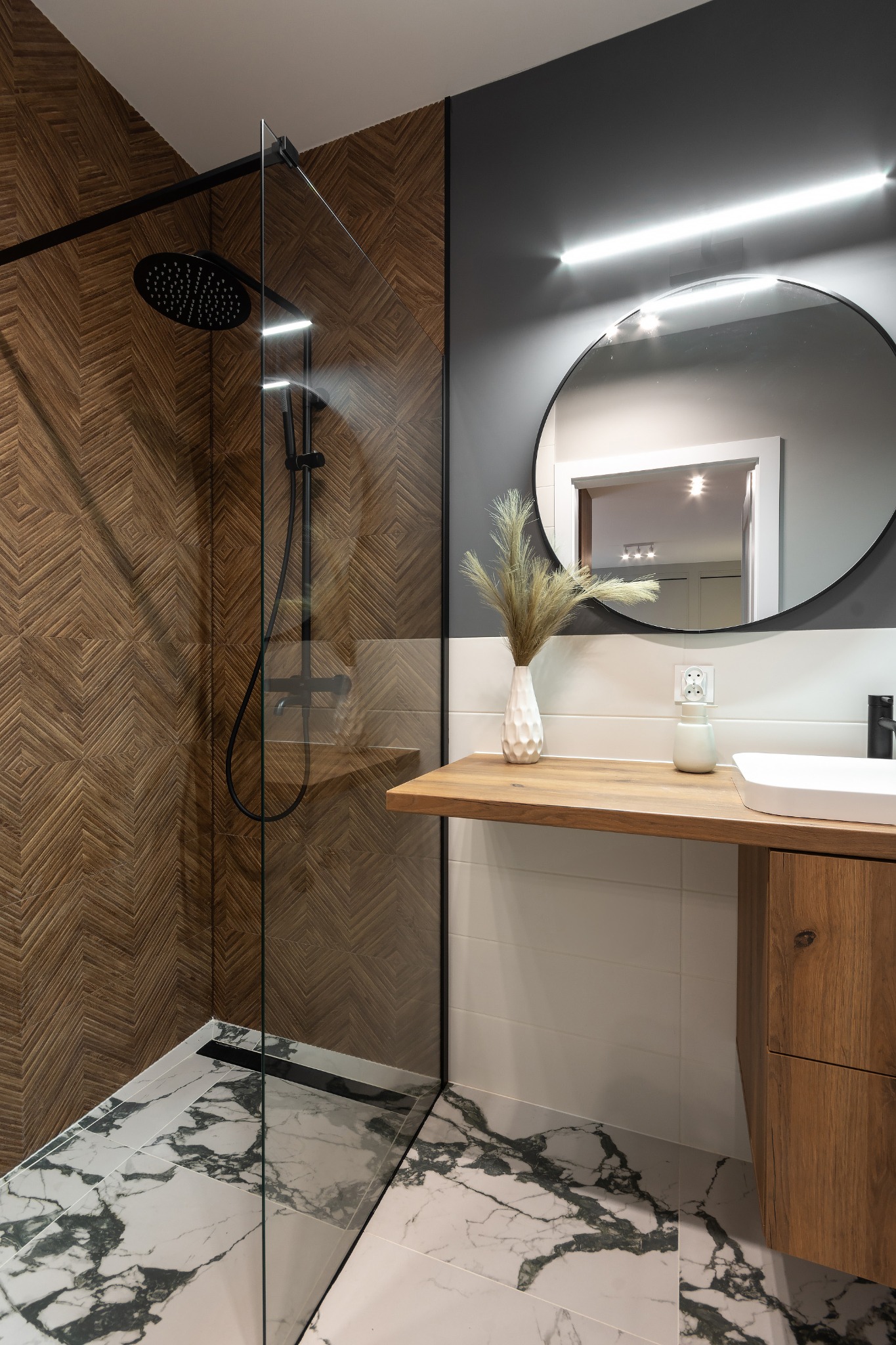 Nowoczesna łazienka z kabiną prysznicową typu walk-in, wykończona drewnopodobną okładziną w geometryczny wzór i marmurową posadzką, okrągłe lustro z oświetleniem LED nad drewnianym blatem z umywalką.