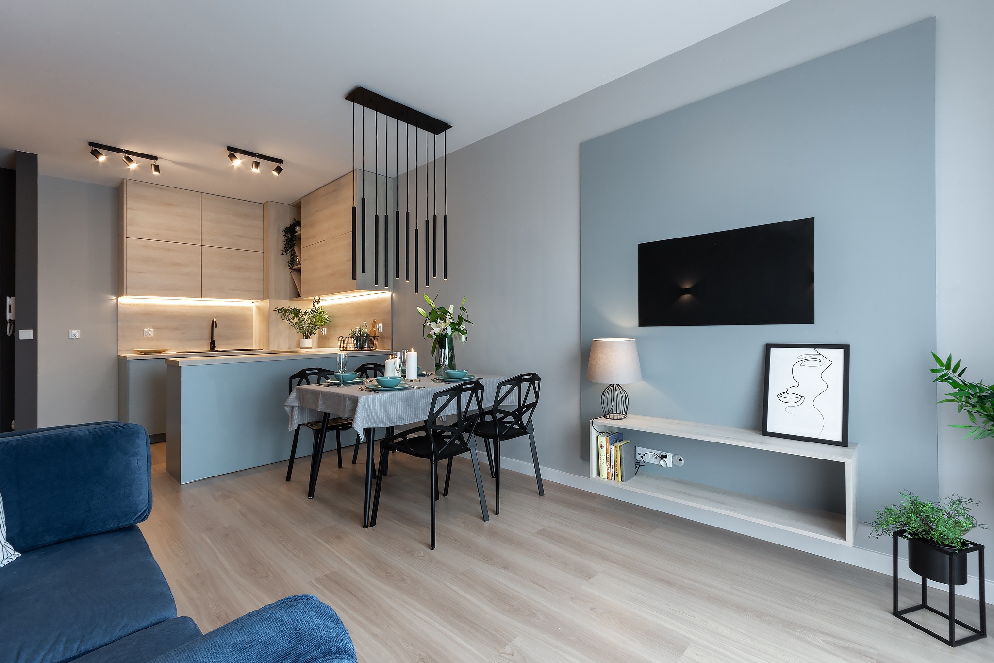 Nowoczesne wnętrze mieszkania w Łodzi: otwarta kuchnia z drewnianymi szafkami, stół jadalny z czarnymi krzesłami i dekoracyjnym oświetleniem, minimalistyczny salon z telewizorem i roślinami.