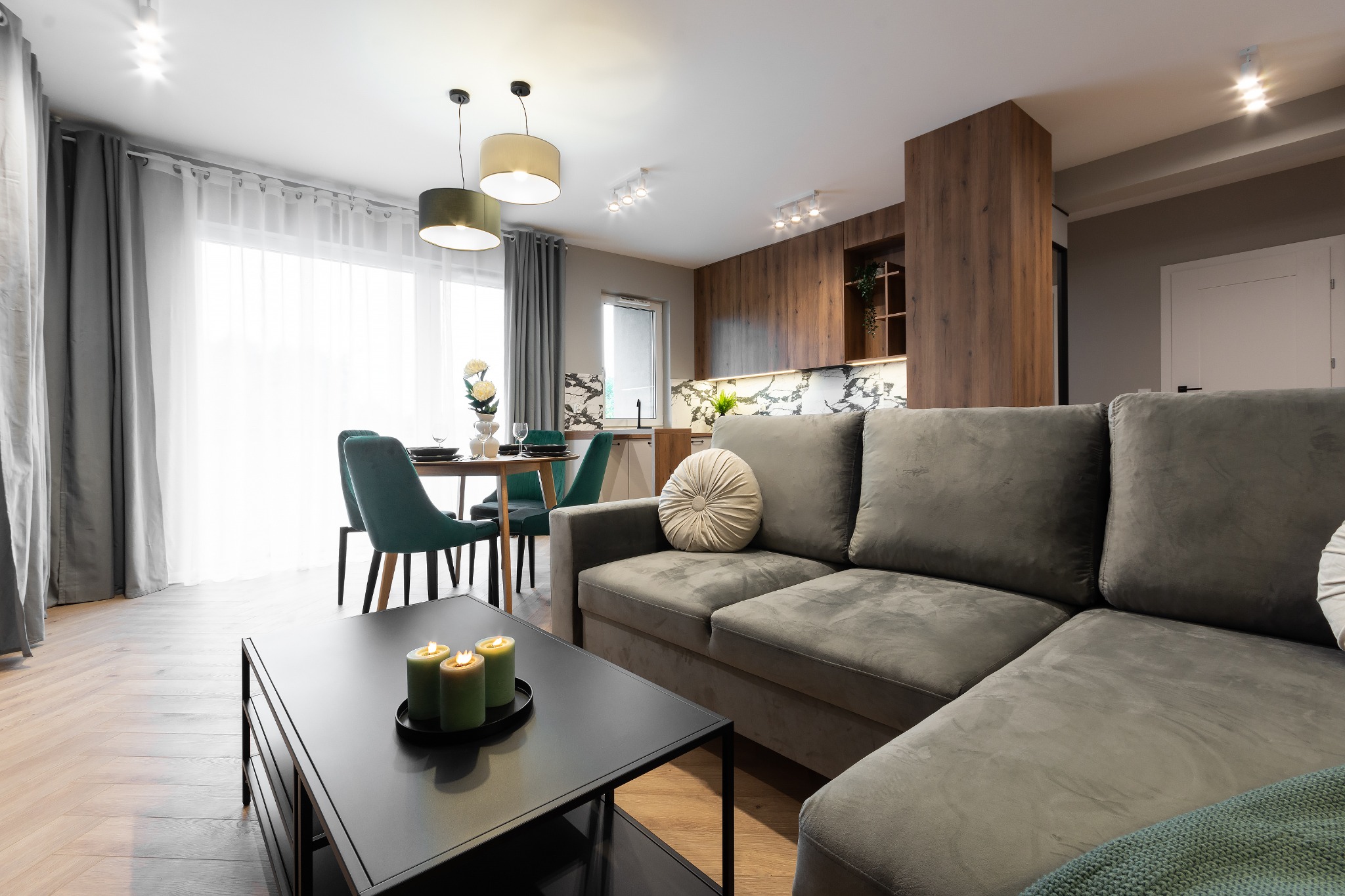 Nowoczesny salon z szarym narożnikiem, czarnym stolikiem kawowym z zapalonymi zielonymi świecami, jadalnią z turkusowymi krzesłami i kuchnią z drewnianymi szafkami w tle.