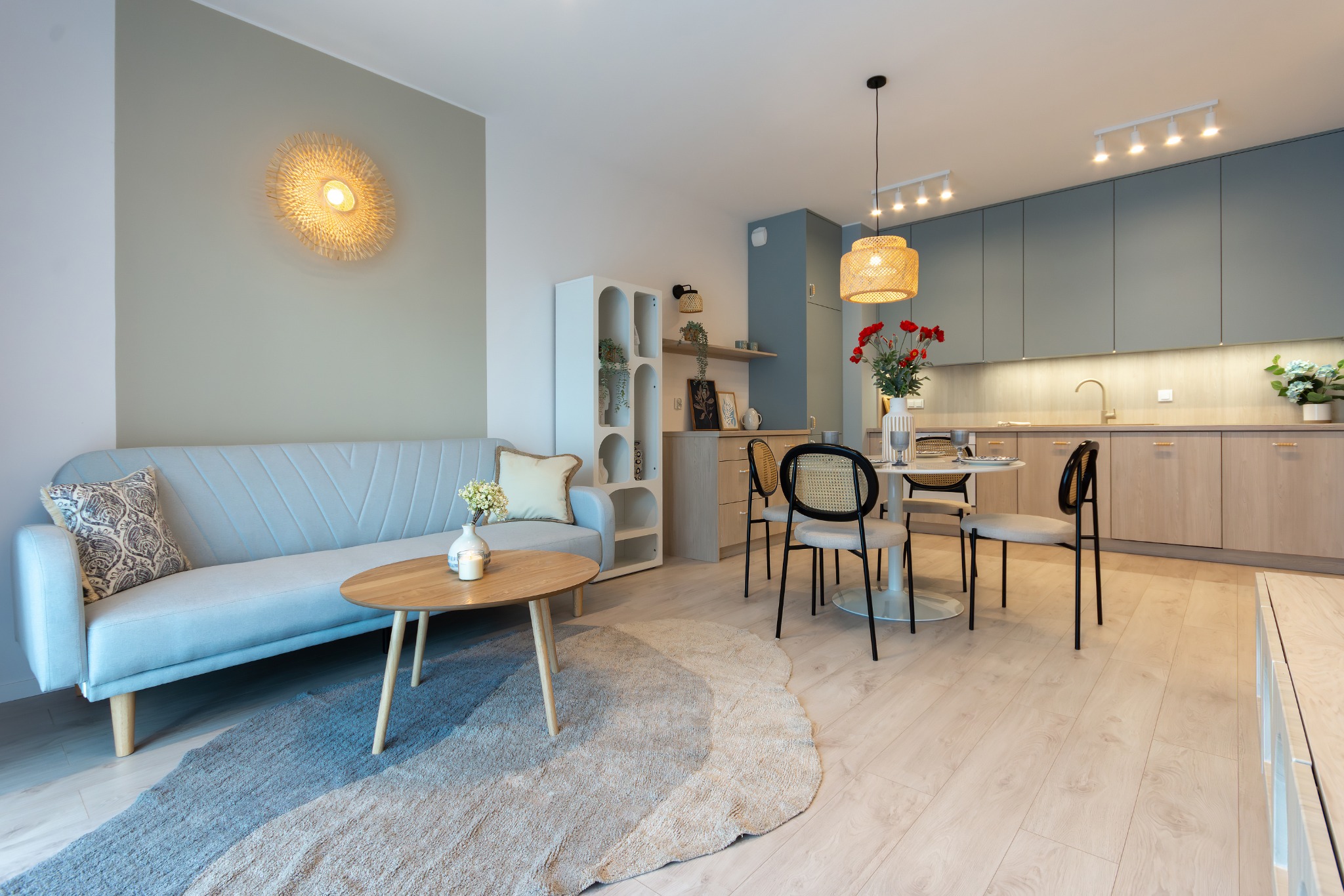 Jasne, przestronne wnętrze mieszkania z sofą, okrągłym stolikiem kawowym na dywanie, stołem z krzesłami i aneksem kuchennym z drewnianymi szafkami oraz dekoracyjnym oświetleniem.