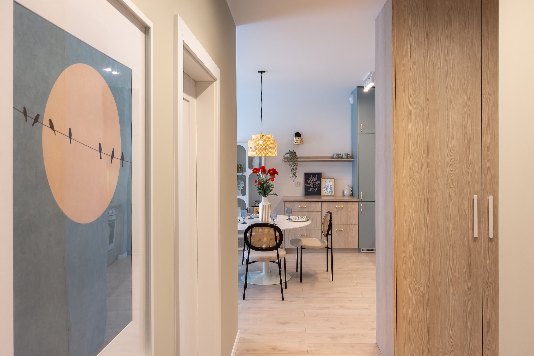 Nowoczesne wnętrze mieszkania z widokiem na okrągły stół jadalny, krzesła z rattanu, kwiaty w wazonie, drewniane szafki kuchenne i minimalistyczny obraz z motywem ptaków.