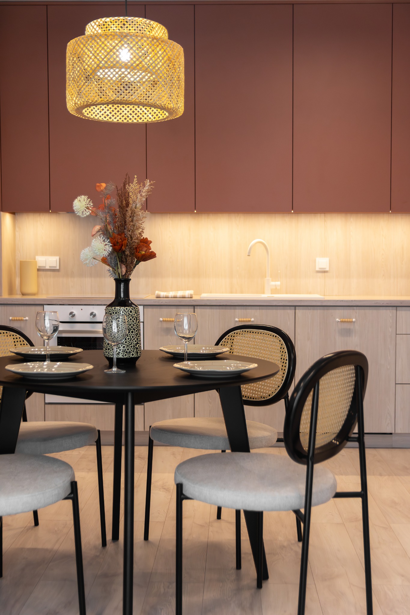 Jadalnia z okrągłym czarnym stołem, nakrytym dla czterech osób, rattanowe krzesła, dekoracyjny wazon z suszonymi kwiatami i pleciona lampa wisząca na tle kuchni z drewnianymi szafkami i różowymi...
