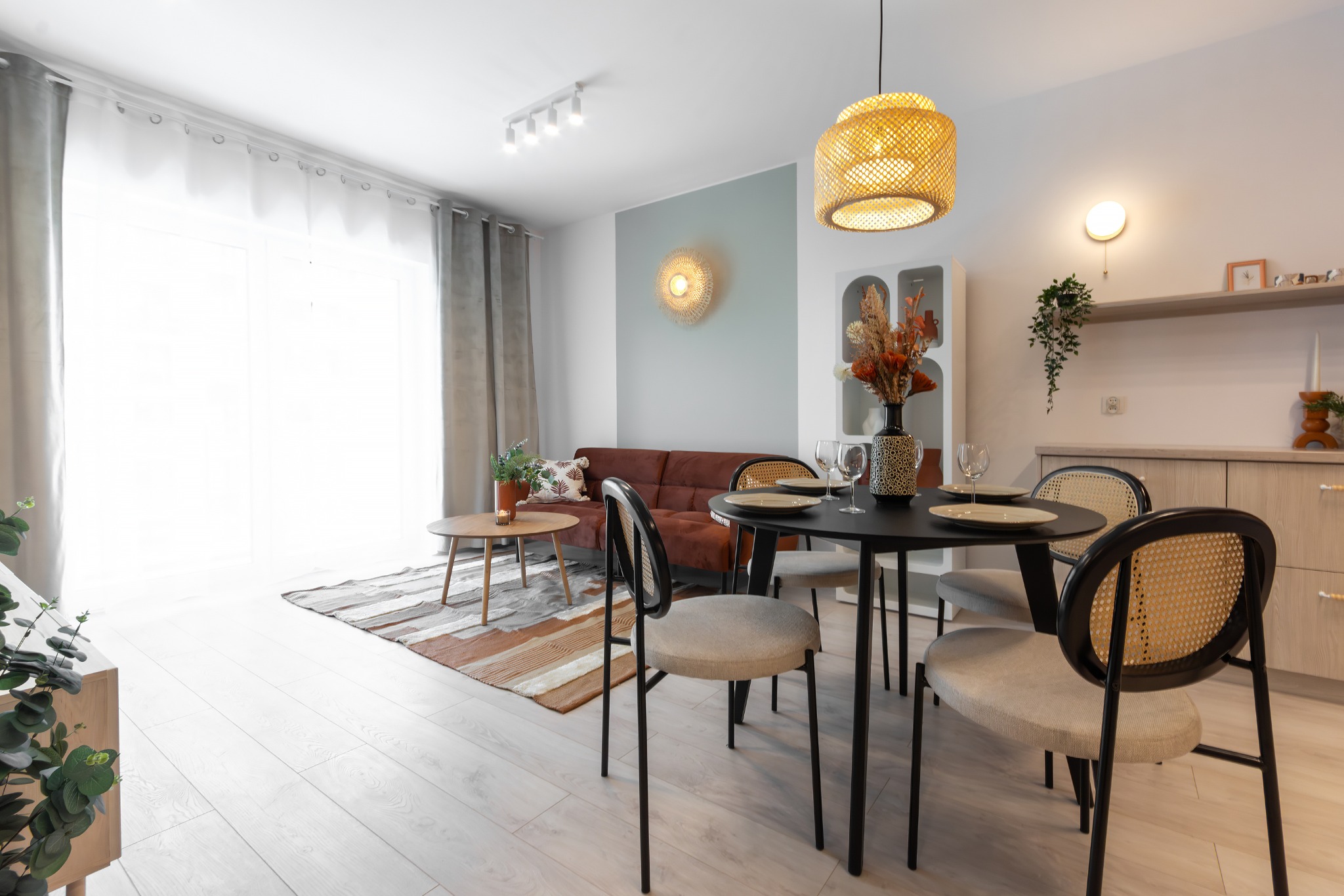 Jasne wnętrze mieszkania z brązową sofą, okrągłym stołem z czarnym blatem i krzesłami z rattanowym oparciem, oświetlone wiszącą lampą z abażurem z plecionki.