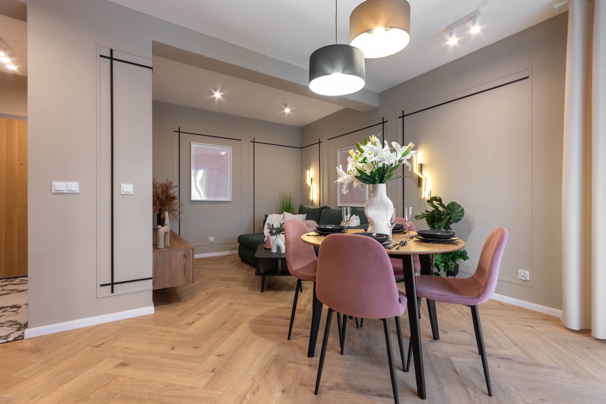 Jasne wnętrze z parkietem w jodełkę, okrągłym stołem z różowymi krzesłami, kwiatami w wazonie i widokiem na salon z zieloną sofą.