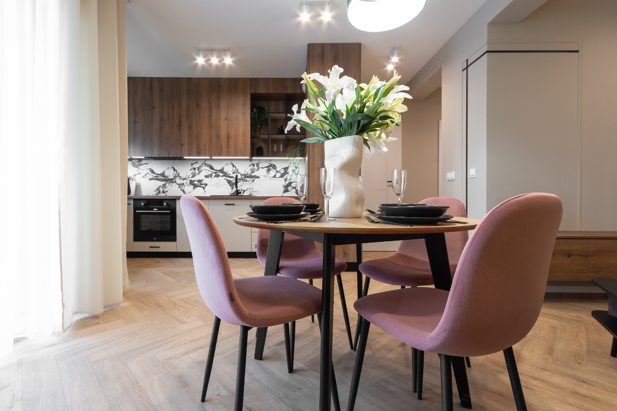 Nowoczesna jadalnia z okrągłym stołem, czterema różowymi krzesłami, białym wazonem z liliami i zastawą stołową na tle otwartej kuchni z drewnianymi szafkami i marmurowym blatem.