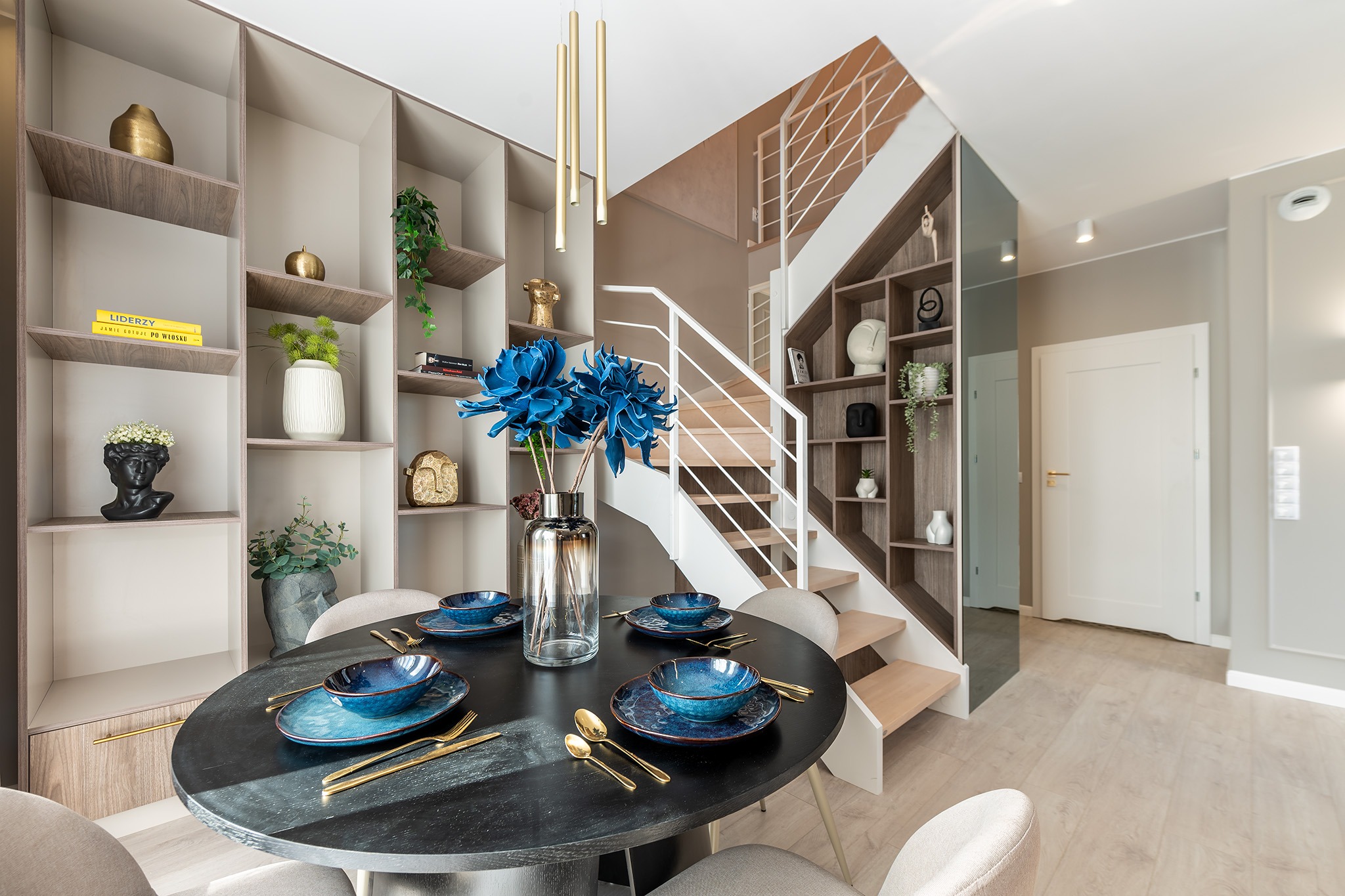 Nowoczesna jadalnia z okrągłym, czarnym stołem, niebieską zastawą i złotymi sztućcami, na tle schodów z zabudową regałową i dekoracjami.