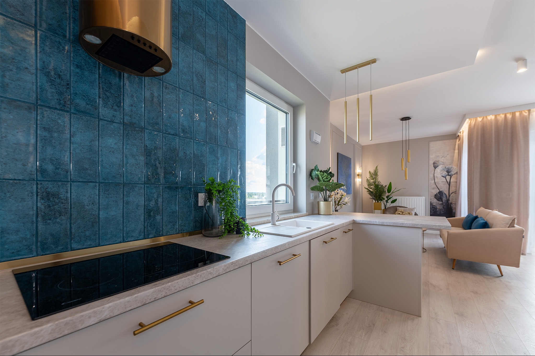 Nowoczesna kuchnia z niebieskimi płytkami imitującymi cegłę, złotym okapem i elementami wykończenia, jasnymi szafkami z podłużnymi złotymi uchwytami i widokiem na salon.
