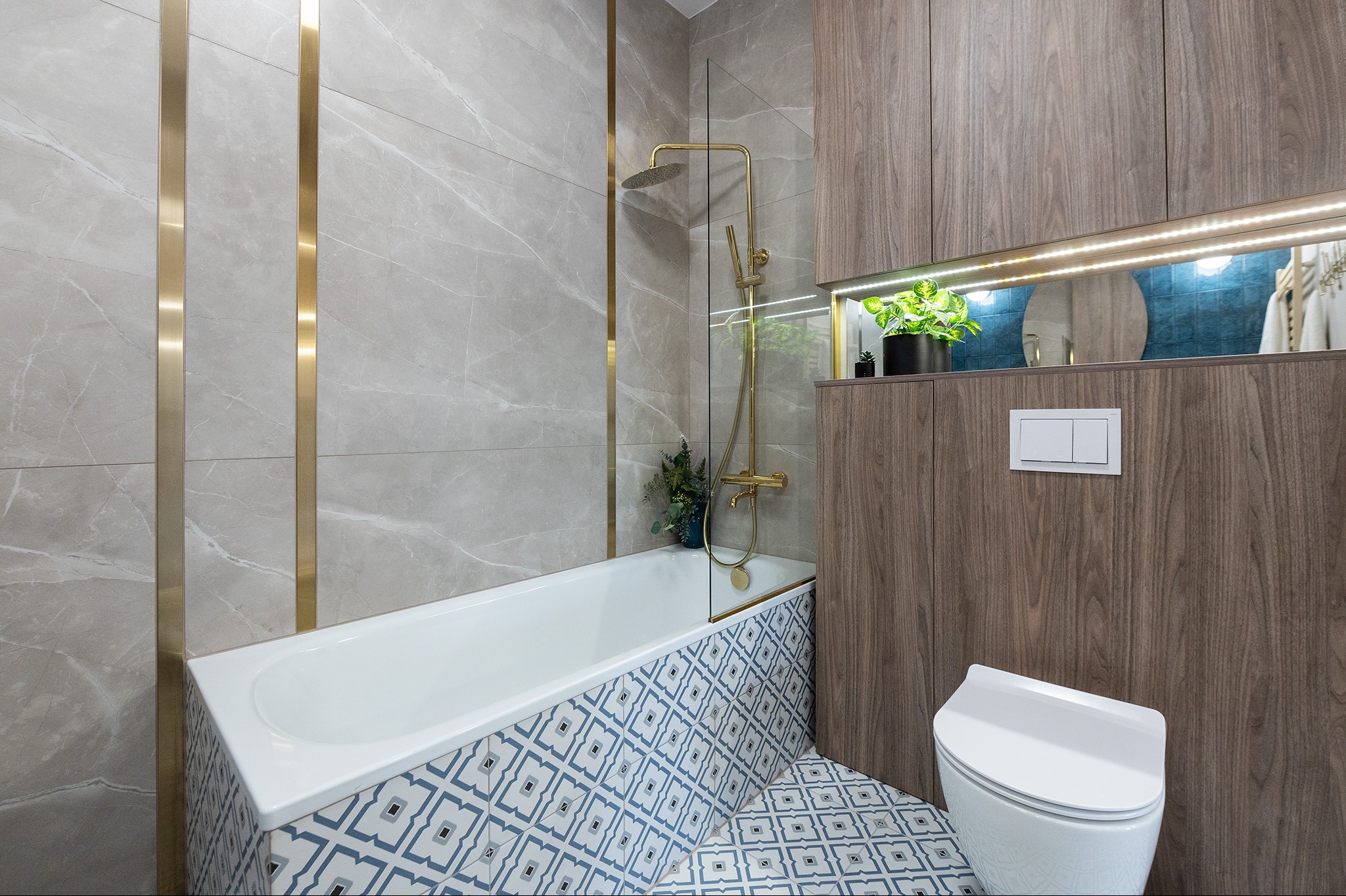 Nowoczesna łazienka z wanną obudowaną w biało-niebieskie płytki, złotą armaturą, szarymi marmurowymi płytkami na ścianach z dekoracyjnymi złotymi listwami oraz zabudową meblową z drewnopodobnego...
