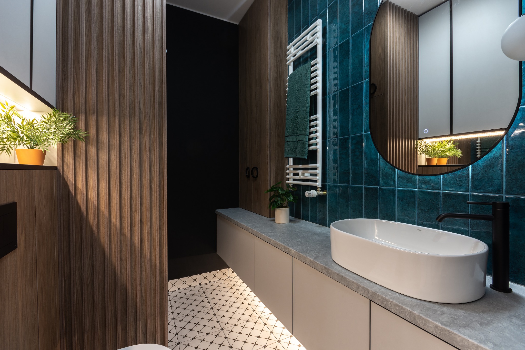 Nowoczesna łazienka z turkusowymi płytkami, owalną umywalką nablatową i grzejnikiem drabinkowym, akcenty drewna i roślin doniczkowych, podświetlana podłoga z wzorem.