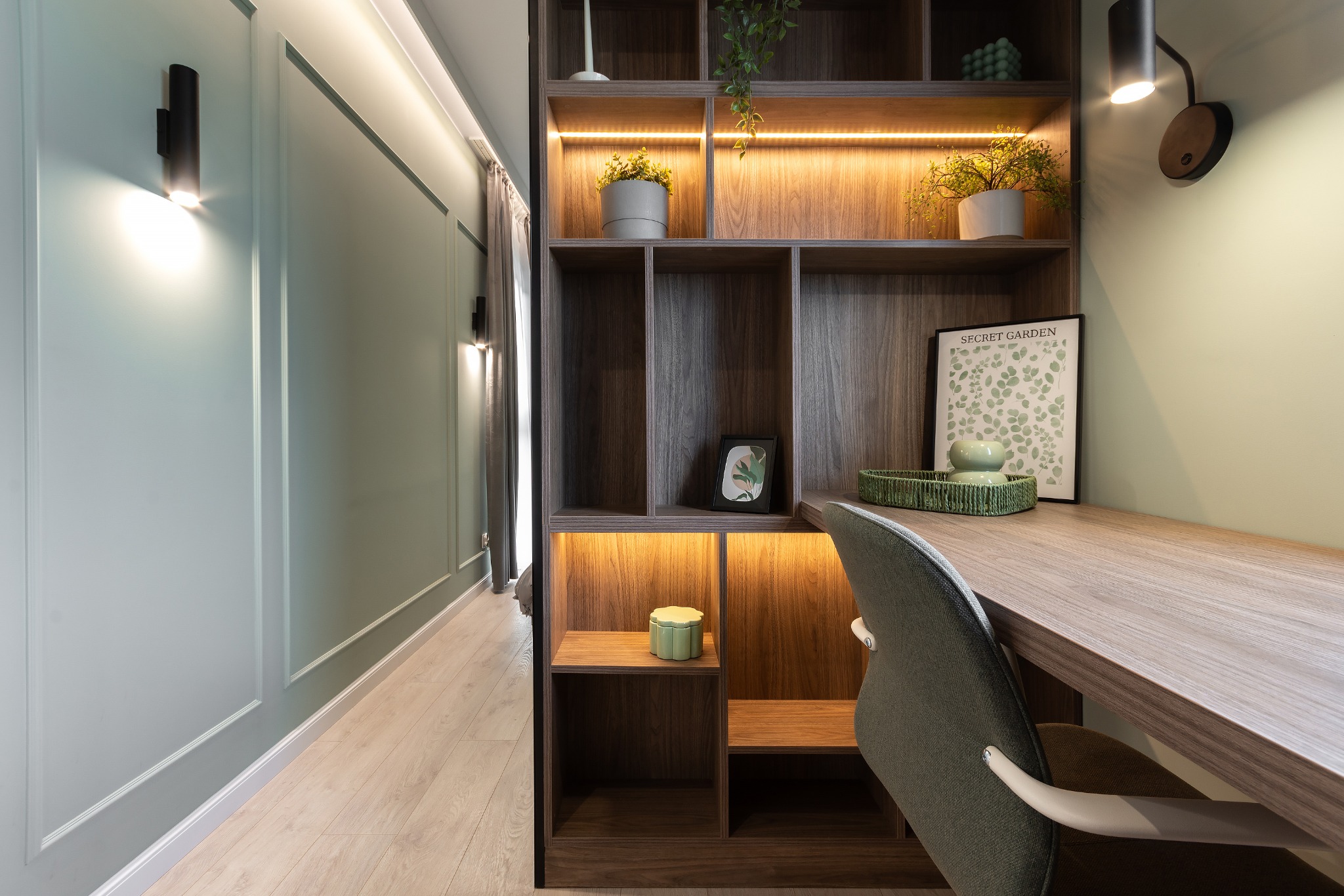 Biurko z wbudowanym regałem oświetlonym od wewnątrz, zielona ściana z dekoracyjnymi listwami, drewniana podłoga, nowoczesny styl.