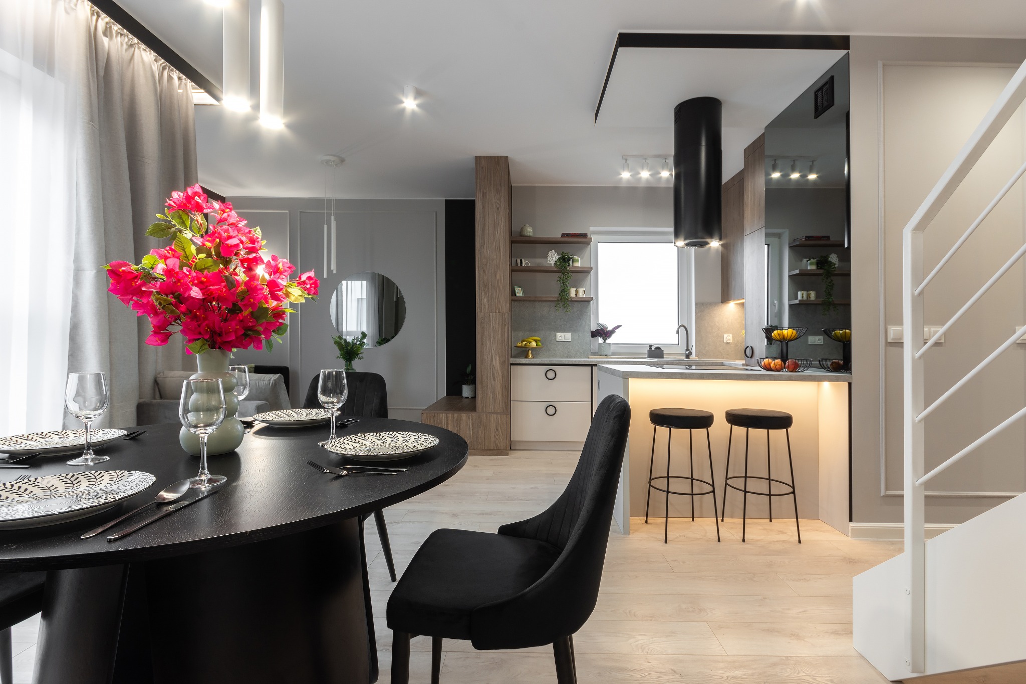 Nowoczesne wnętrze jadalni z czarnym okrągłym stołem, nakrytym do posiłku, z bukietem różowych kwiatów, połączonej z otwartą kuchnią z wyspą i hokerami, oraz białymi schodami.