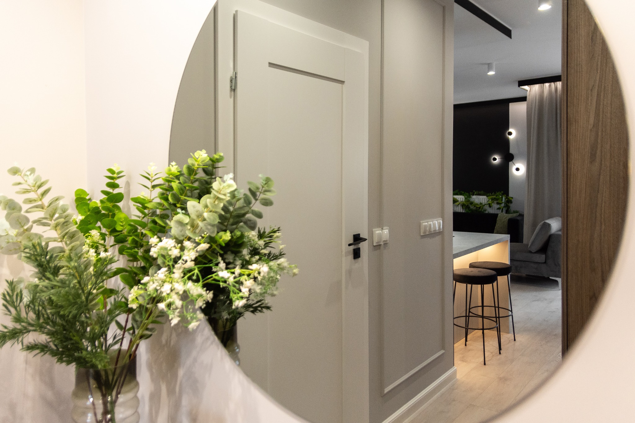 Okrągłe lustro odbija fragment nowoczesnego wnętrza: szarą ścianę z białą listwą przypodłogową, białe drzwi z czarną klamką, oraz widok na salon z dwoma czarnymi stołkami barowymi i szarą sofą.