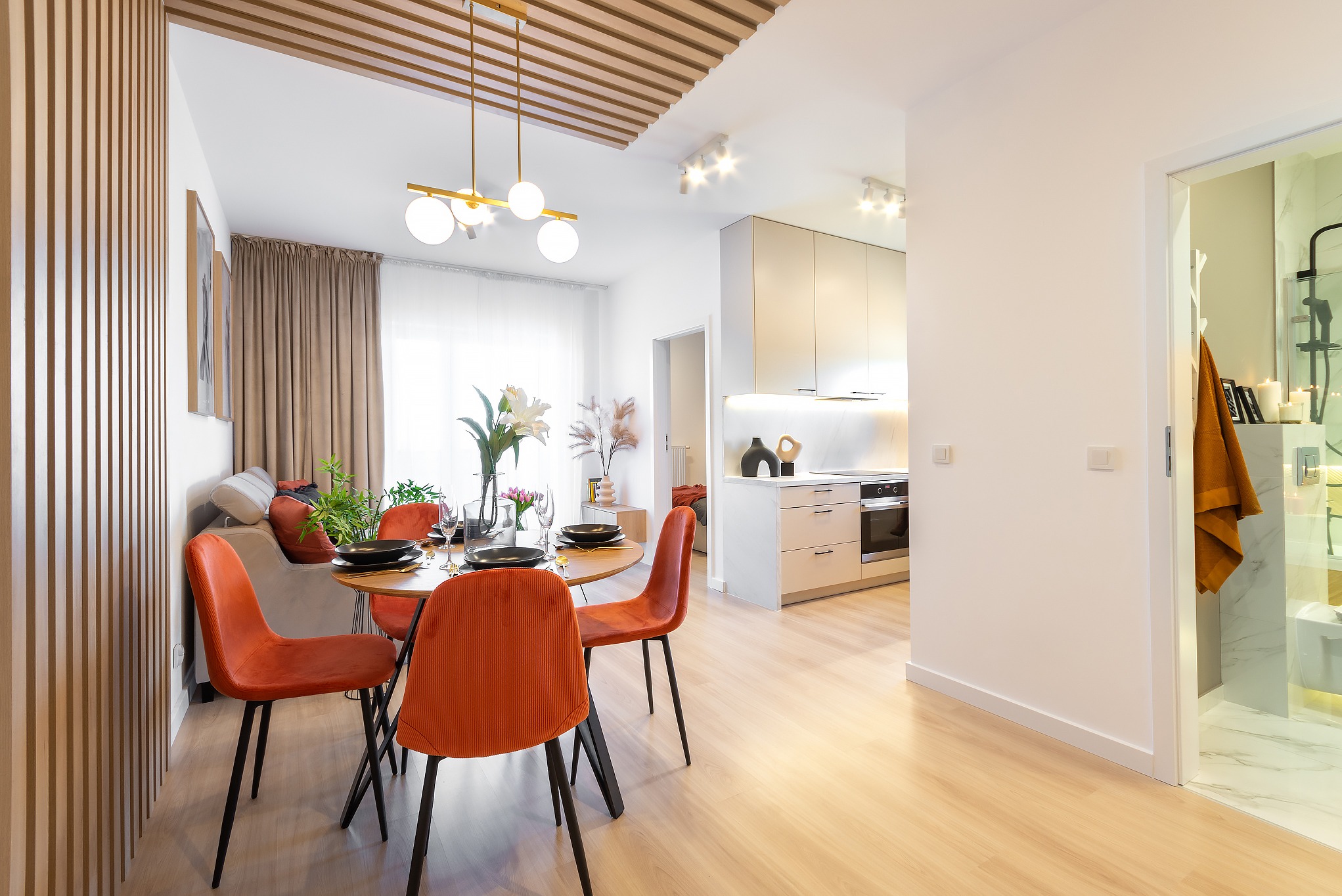 Jasne wnętrze mieszkania z okrągłym stołem i pomarańczowymi krzesłami, widok na kuchnię i łazienkę z kabiną prysznicową.