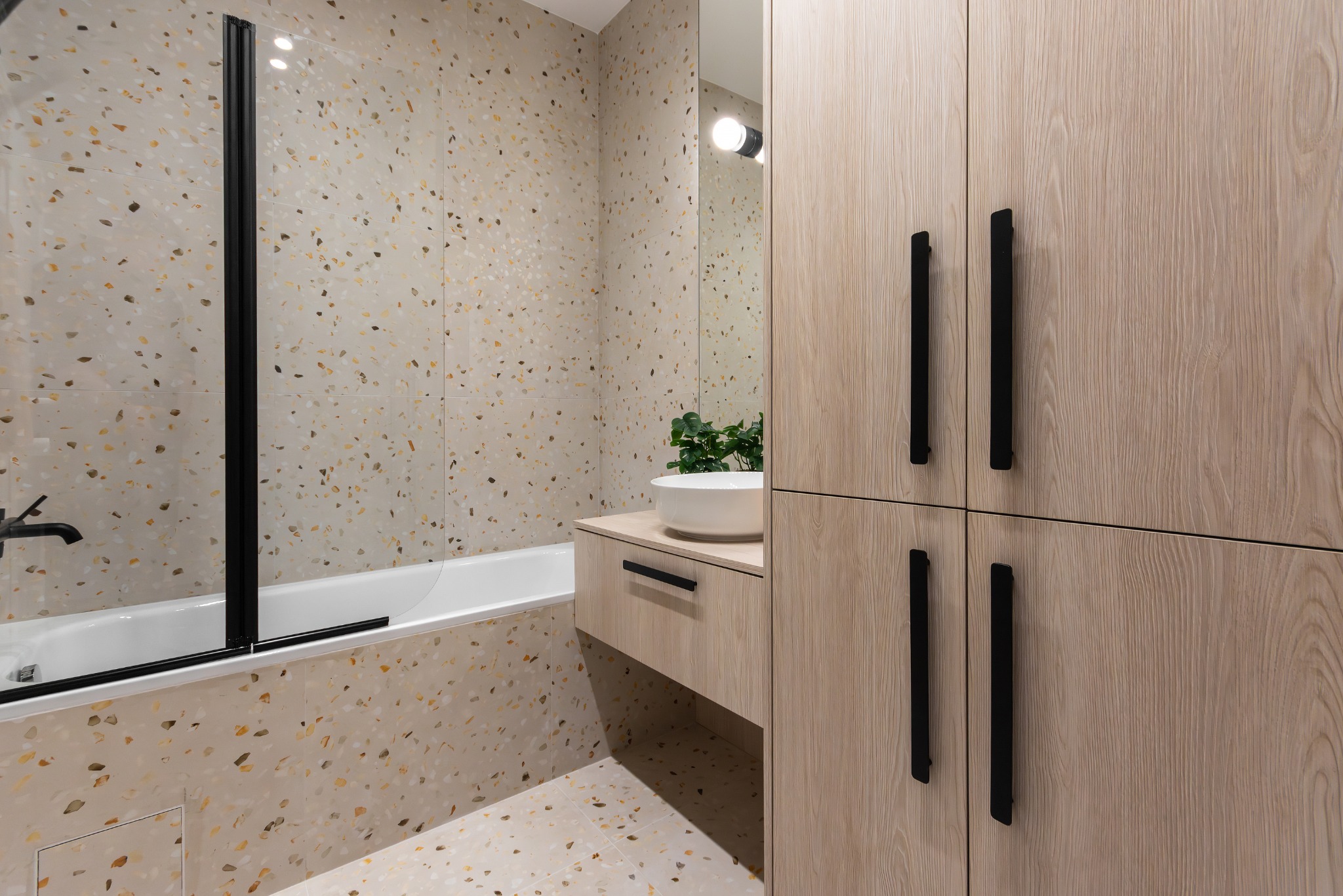 Nowoczesna łazienka z terazzo, białą umywalką nablatową, szafką z drewnopodobnymi frontami i czarnymi uchwytami oraz czarną armaturą przy wannie.