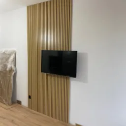 Drewniane lamele ścienne za telewizorem, widoczne fragmenty podłogi, folia ochronna na meblu w rogu, gniazdko elektryczne w listwie.