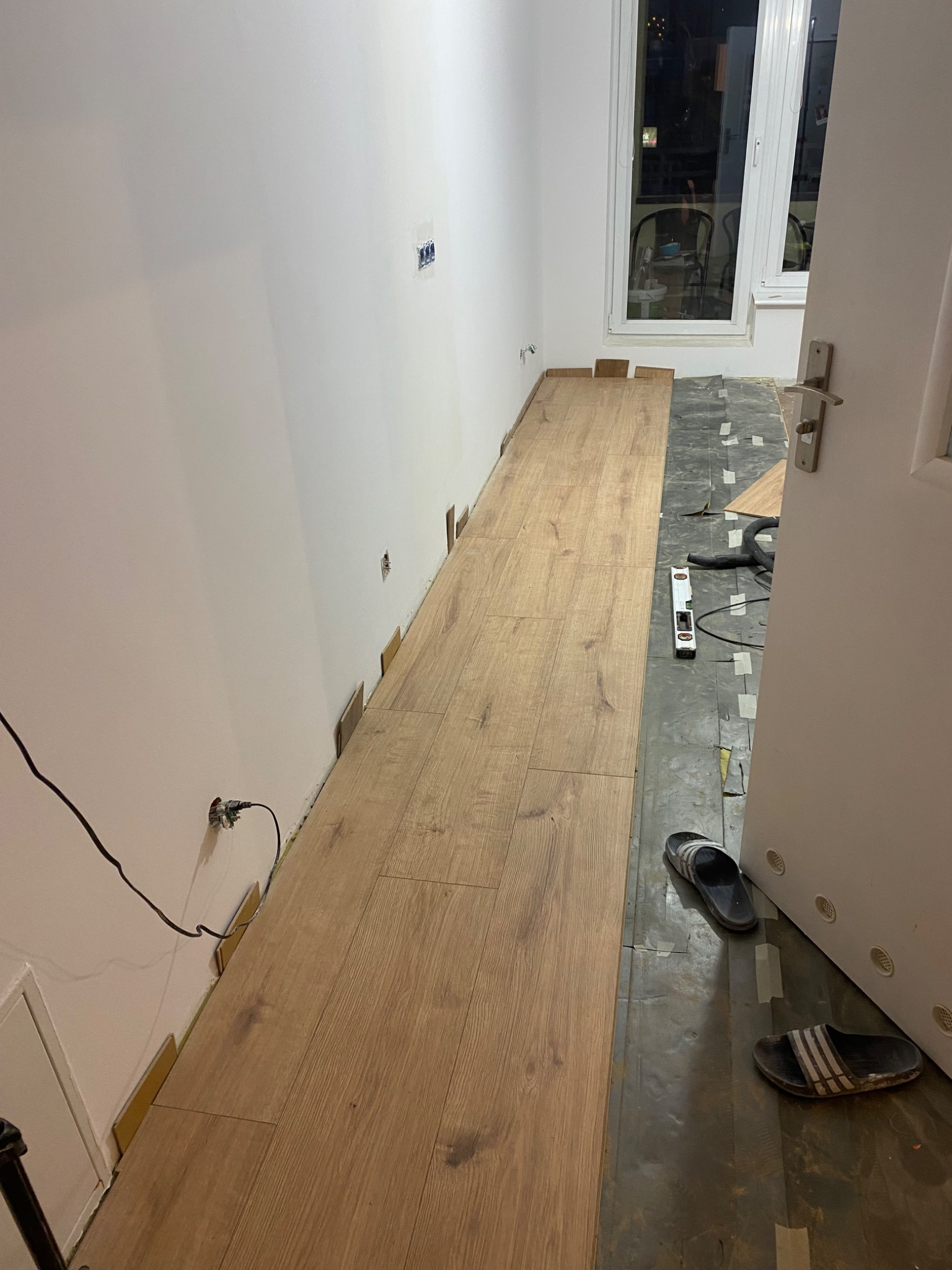 Układanie paneli podłogowych w wąskim pomieszczeniu, widoczne niedopasowane panele przy ścianie, poziomica, odkurzacz i klapki na podłodze.