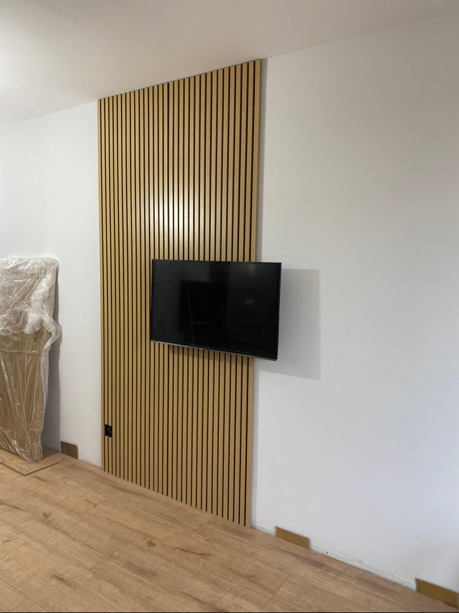 Drewniane lamele ścienne za telewizorem, widoczne fragmenty podłogi, folia ochronna na meblu w rogu, gniazdko elektryczne w listwie.