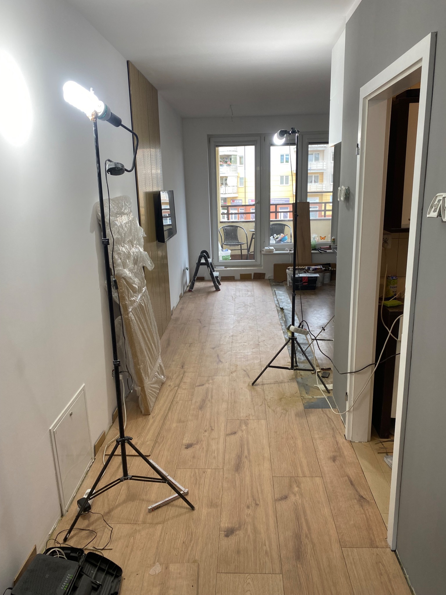 Wnętrze mieszkania w trakcie układania paneli podłogowych, widoczne dwie lampy budowlane na trójnogach, panele pod ścianą w folii, częściowo ułożona podłoga z paneli, narzędzia i akcesoria...