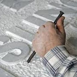 PRACOWNIA KAMIENIA LEXBUD SP Z O.O. - Ręka w koszuli w kratę dłutuje litery w jasnym kamieniu za pomocą dłuta i młotka, tworząc napis o nieznanej treści.