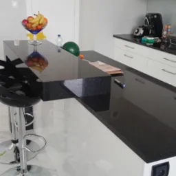 Czarny, błyszczący blat kuchenny z konglomeratu z owocami w szklanej misie, butelką wody, balonem i czarnymi hokerami na chromowanej nodze w nowoczesnej kuchni.