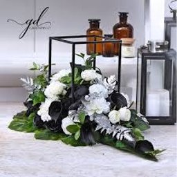 PRACOWNIA KAMIENIA LEXBUD SP Z O.O. - Dekoracja nagrobna z białych i czarnych kwiatów, srebrnymi liśćmi i zielonymi elementami, umieszczona na jasnym blacie, z metalowym, czarnym kubikiem w tle i brązowymi flakonami.