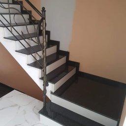 PRACOWNIA KAMIENIA LEXBUD SP Z O.O. - Schody wewnętrzne z czarnego kamienia z ozdobną metalową balustradą, białe podstopnice, marmurowa podłoga, ściana w dwóch odcieniach beżu.