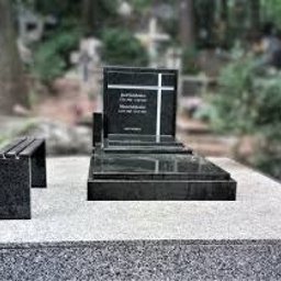 PRACOWNIA KAMIENIA LEXBUD SP Z O.O. - Czarny, granitowy pomnik z wyrytym krzyżem i napisem, ustawiony na jasnej płycie nagrobnej, obok mała ławka z tego samego materiału, w tle zieleń cmentarza.