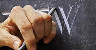 Ręka kamieniarza dłutem wykuwa literę 'W' w ciemnym kamieniu, tworząc napis.