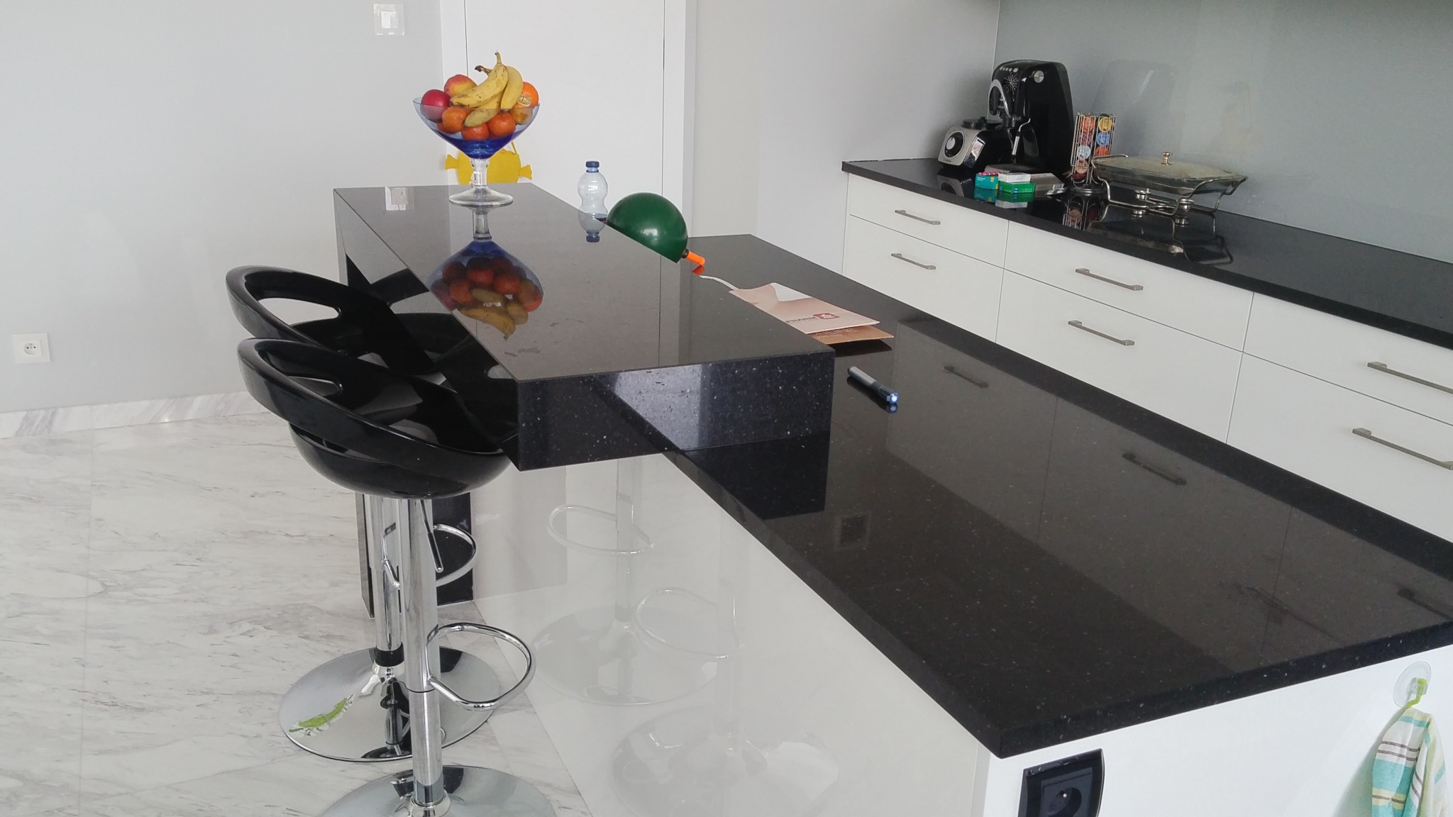 Czarny, błyszczący blat kuchenny z konglomeratu z owocami w szklanej misie, butelką wody, balonem i czarnymi hokerami na chromowanej nodze w nowoczesnej kuchni.