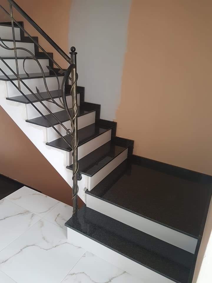 Schody wewnętrzne z czarnego kamienia z ozdobną metalową balustradą, białe podstopnice, marmurowa podłoga, ściana w dwóch odcieniach beżu.