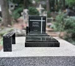 Czarny, granitowy pomnik z wyrytym krzyżem i napisem, ustawiony na jasnej płycie nagrobnej, obok mała ławka z tego samego materiału, w tle zieleń cmentarza.