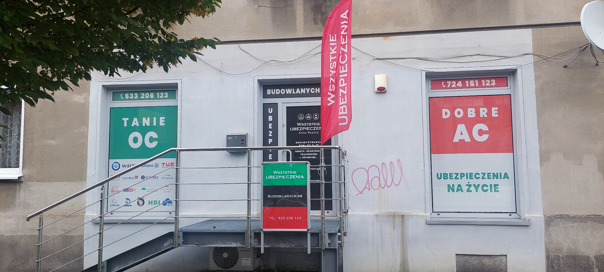 Fasada biura ubezpieczeń z reklamami 'Tanie OC', 'Dobre AC', 'Wszystkie ubezpieczenia', flaga reklamowa i schody wejściowe. Widoczne numery telefonów i nazwa firmy.