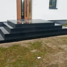 Schody wejściowe wyłożone czarną, błyszczącą kostką granitową, prowadzące do jasnego domu z drewnianymi drzwiami.