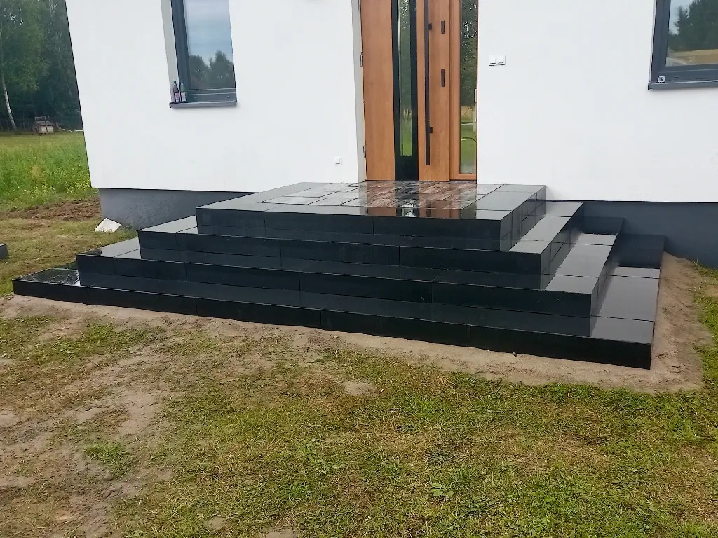 Nowoczesne, czarne schody wejściowe z błyszczącej kostki prowadzące do jasnego domu z drewnianymi drzwiami, widoczne trawnik i fragment okna.