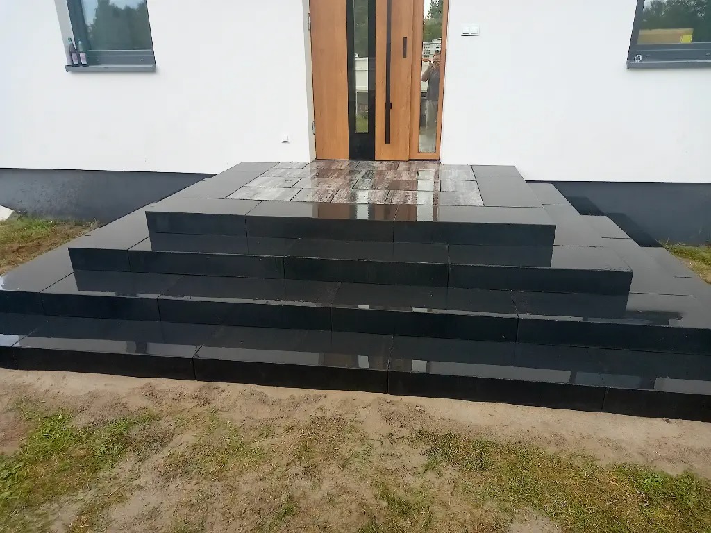 Schody wejściowe do domu wyłożone czarną, błyszczącą kostką granitową, z widocznymi refleksami światła na mokrej powierzchni.