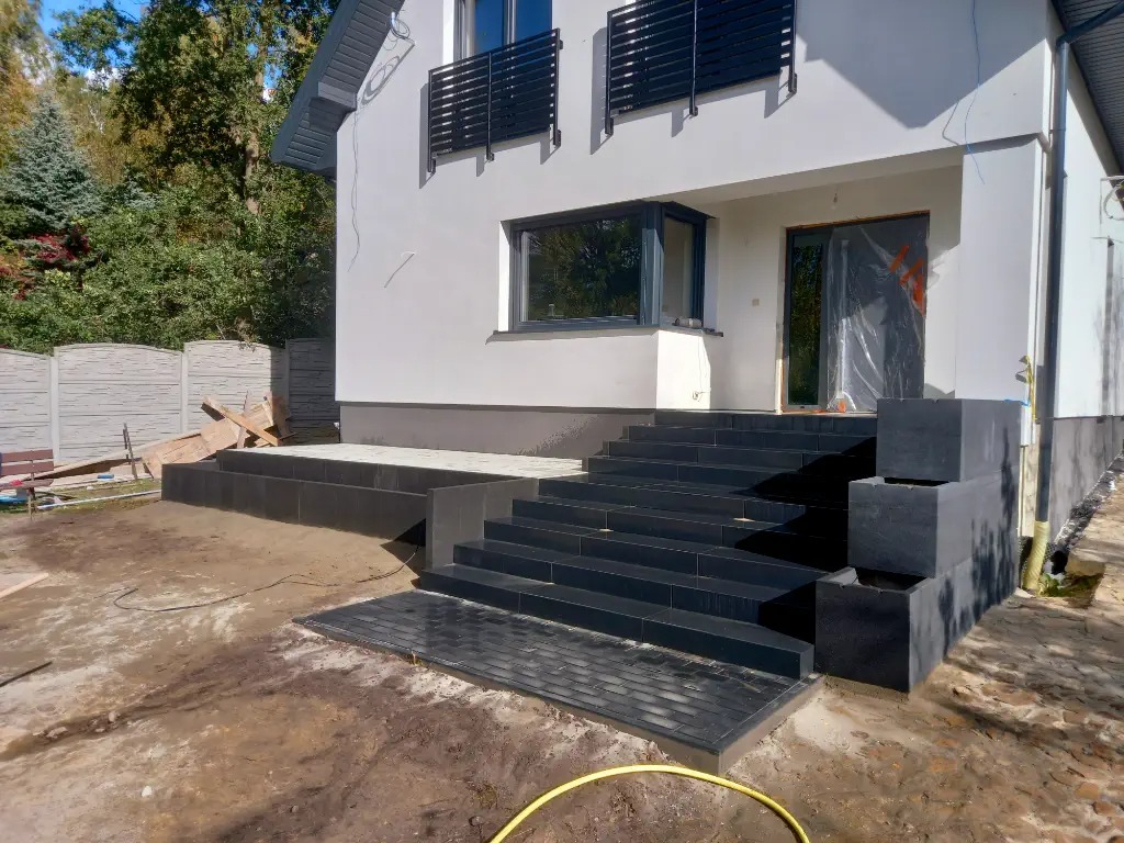 Schody wejściowe do domu wyłożone ciemną kostką, z widocznym fragmentem żółtego węża ogrodowego na pierwszym planie.