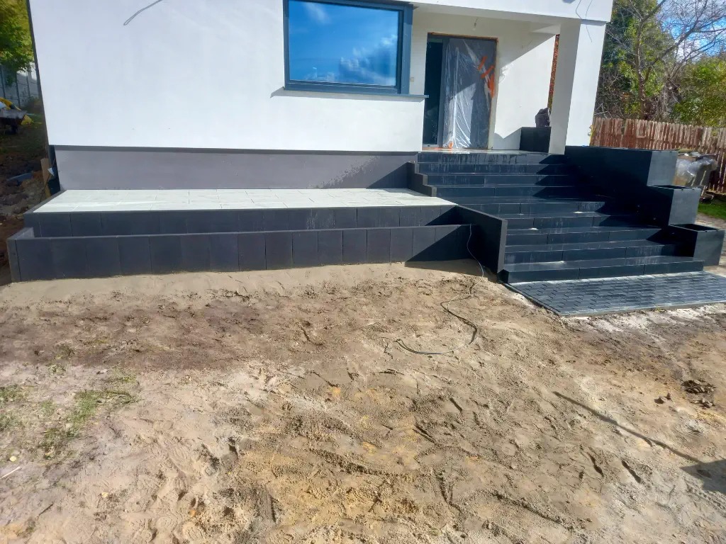 Nowoczesny taras z ciemnej kostki brukowej prowadzący do domu z białymi ścianami, widoczne schody i fragment terenu przed budynkiem w trakcie prac wykończeniowych.