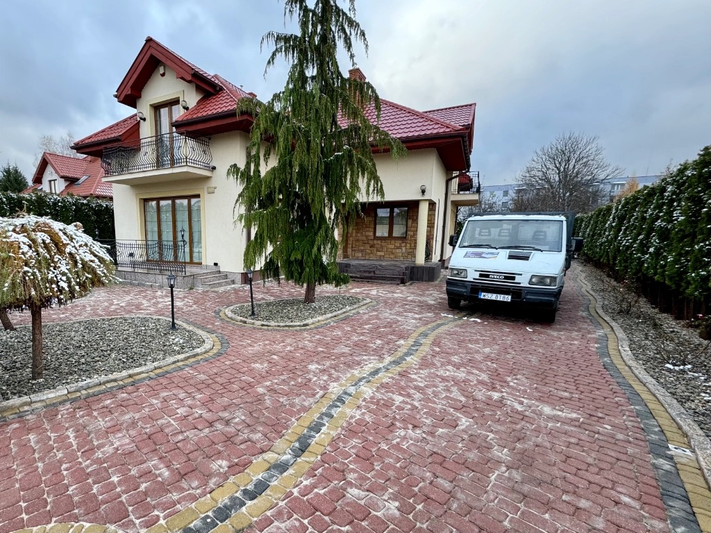 Czerwona kostka brukowa na podjeździe do domu z białego kamienia, z czarno-żółtym obramowaniem, częściowo pokryta śniegiem, z zaparkowanym białym samochodem dostawczym.