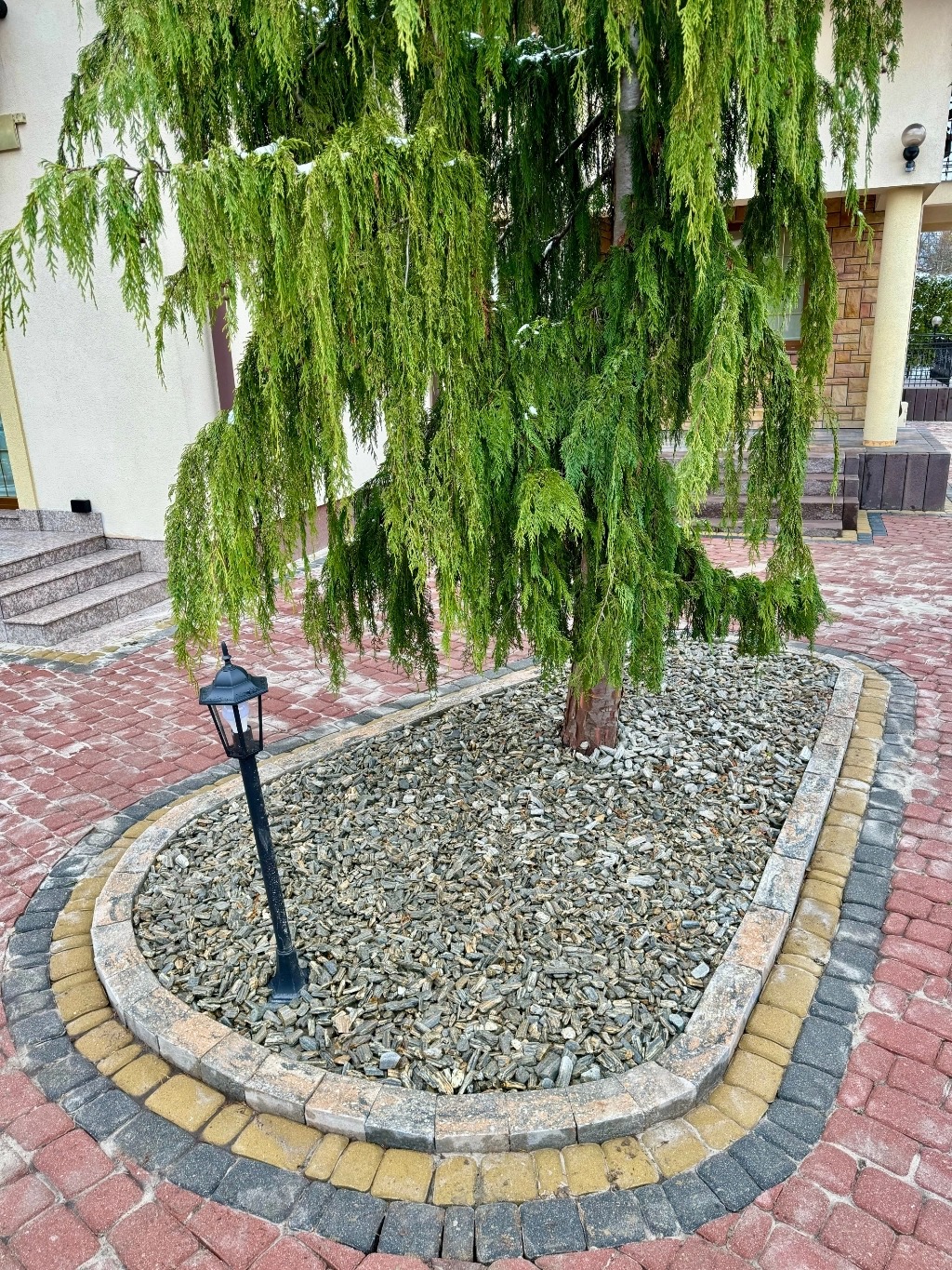 Obrzeże z kostki brukowej wokół drzewa iglastego, wypełnione kora kamienną, z lampą ogrodową w tle domu.