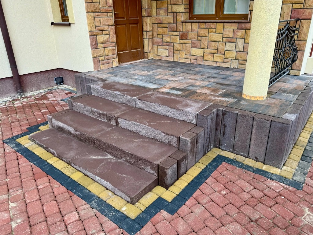 Schody wejściowe z brązowej kostki, obramowane żółto-czarną kostką, prowadzące na taras z szarej kostki, na tle elewacji z kamienia piaskowca.
