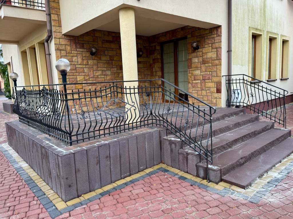 Wejście do domu z brukowanymi schodami i ozdobną, kutą balustradą, obrzeża z kostki brukowej w kolorze żółtym i szarym, czerwona kostka brukowa na podjeździe.