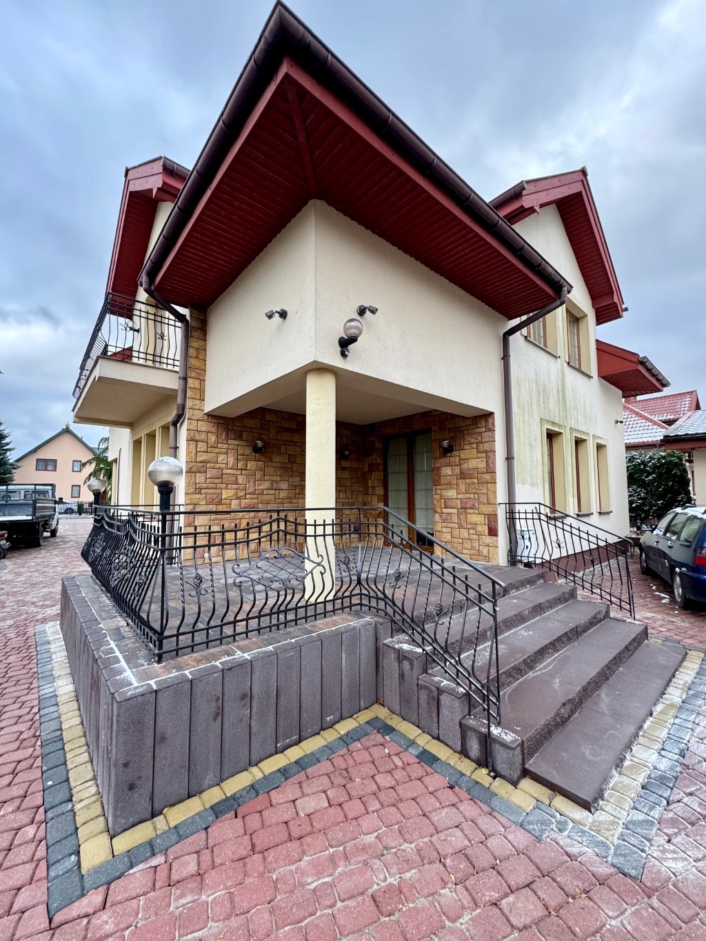 Wejście do domu z kostki brukowej w odcieniach czerwieni i szarości, z ciemnoszarymi schodami i czarną, kutą balustradą, otoczone kostką obrzeżową w kolorze żółtym i szarym.