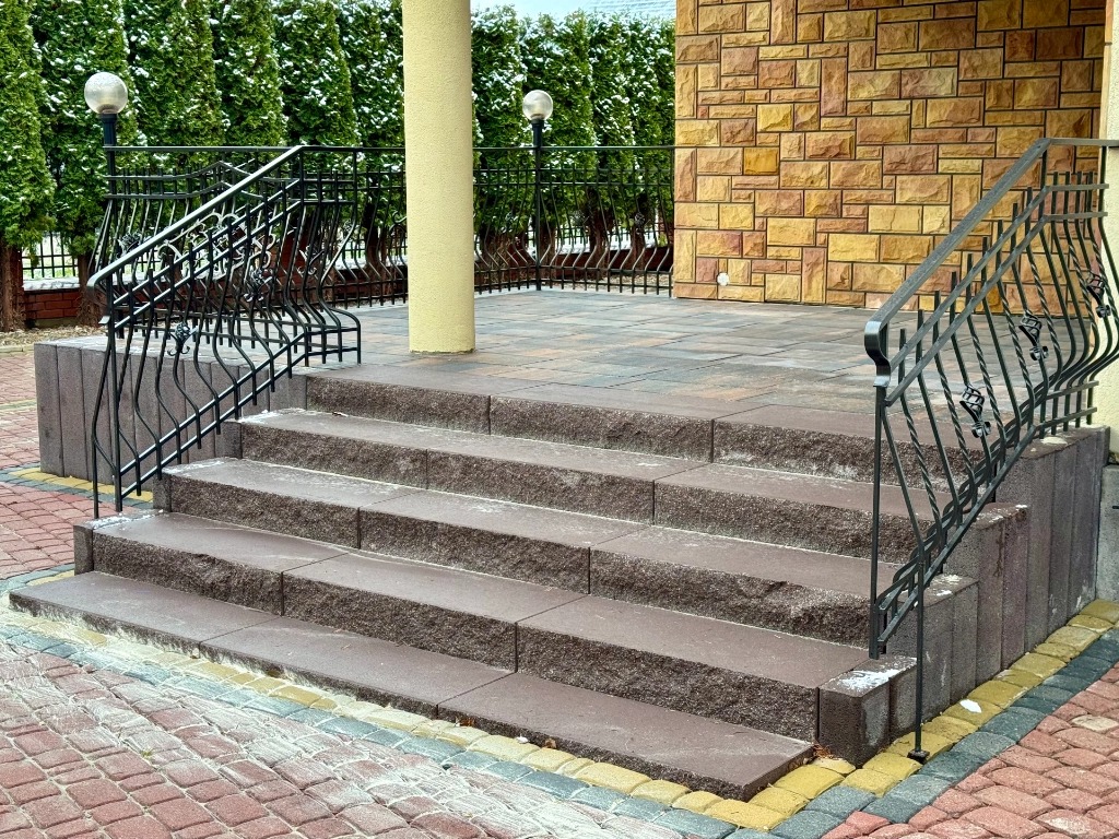 Schody wejściowe wyłożone brązową kostką z ozdobną, kutą balustradą, prowadzące na taras z kostki, z kolumną i elewacją z kamienia dekoracyjnego w tle.
