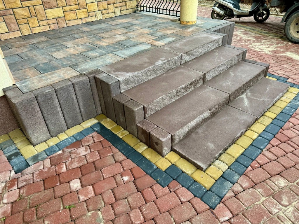Schody wejściowe wyłożone brązową kostką brukową, obrzeża z kostki w kolorach żółtym i grafitowym, w tle fragment elewacji budynku i skuter.