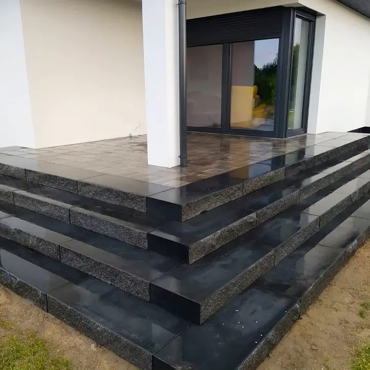 Schody wejściowe do domu wyłożone błyszczącą, czarną kostką granitową, z widoczną strukturą kamienia, prowadzące do przeszklonych drzwi z roletą zewnętrzną.
