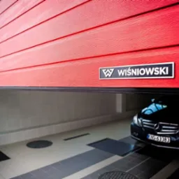 Czerwona segmentowa brama garażowa marki Wiśniowski, lekko uchylona, z widocznym czarnym samochodem Mercedes w garażu z jasnymi kafelkami na ścianach i podłodze.