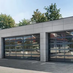 Nowoczesny budynek z dwiema przeszklonymi segmentowymi bramami z szarymi ramami, odbijającymi otoczenie, z widocznym logo producenta Wiśniowski w prawym dolnym rogu.