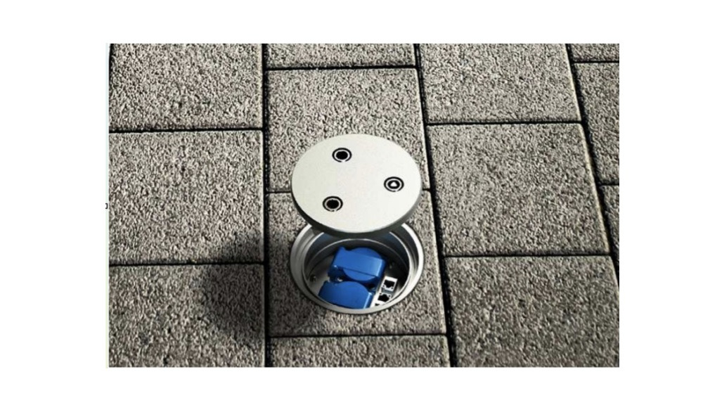 Szara kostka brukowa z aluminiową studzienką rewizyjną z niebieskimi złączkami wewnątrz, widoczna pokrywa z trzema otworami.