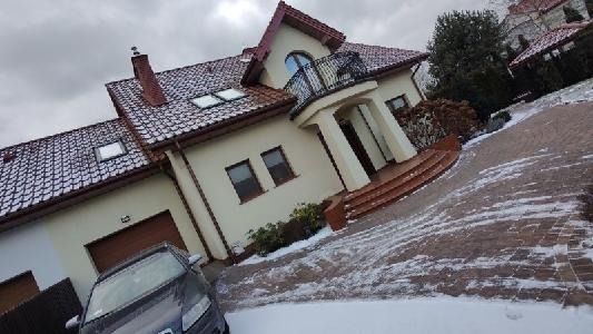 Jasna elewacja domu jednorodzinnego z czerwonym dachem pokrytym śniegiem, widoczny balkon z czarną balustradą, podjazd z kostki brukowej częściowo zasypany śniegiem, pochmurne niebo w tle.