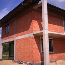 Surowy, dwukondygnacyjny budynek z czerwonej cegły ceramicznej w trakcie budowy, z widocznymi betonowymi słupami i podciągami, ciemne okna wstawione, zwały piasku u podstawy, błękitne niebo...